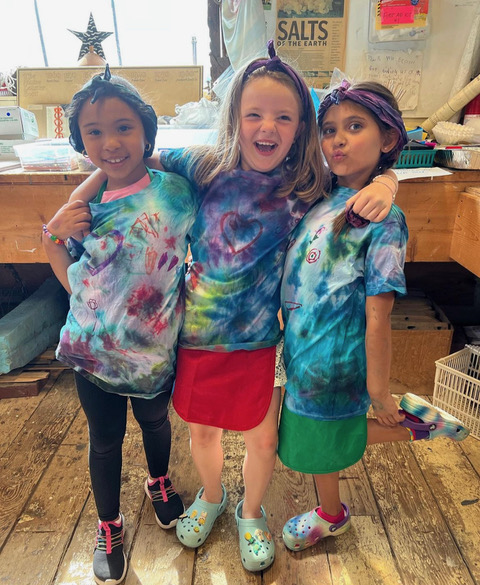 photo of three children standing together, wearing tie-dyed shirts, and smiling at the camera. The girl in the middle has her arm around the both the kid to her right and to her left
