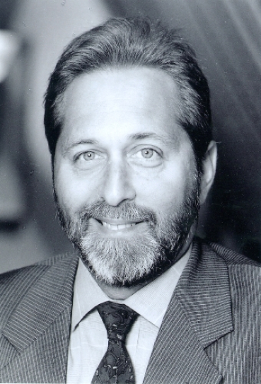 a black-and-white portrait of Lenny Zakim, a white man with a beard, smiling at the camera, wearing a suit jacket, collared shirt, and tie
