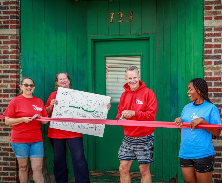 photo of four adults that look to be taking part in a ribbon cutting; three of them have on matching red shirts, one is holding a sign that says '2023 New Home Healthy Waltham'