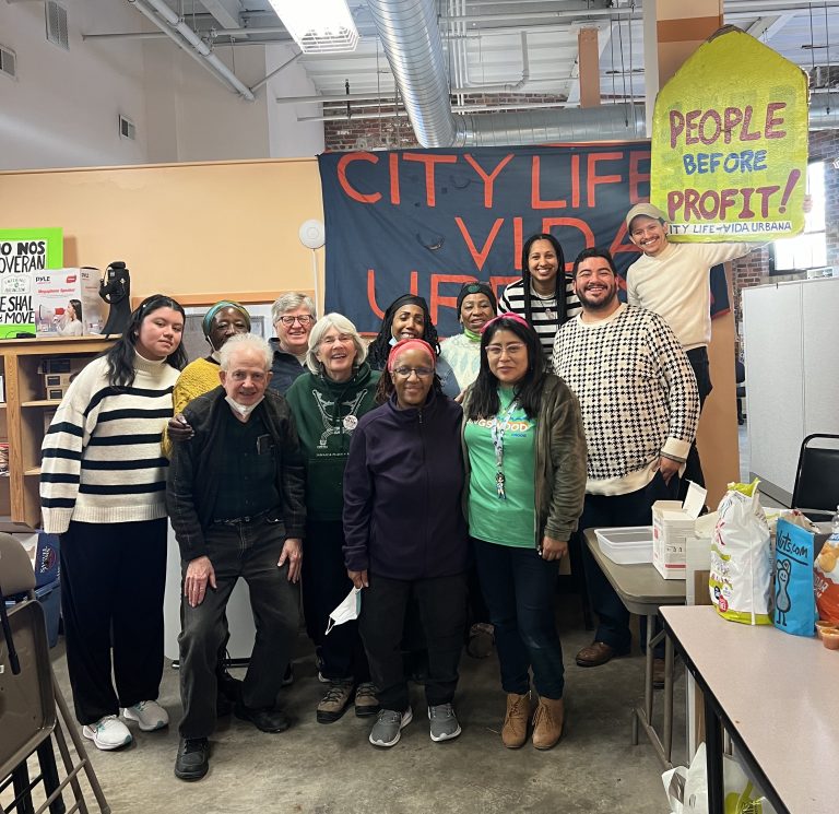 photo of twelve adults standing together, smiling, and posing for the camera in front of a sign that says 'City Life Vida Urbana'