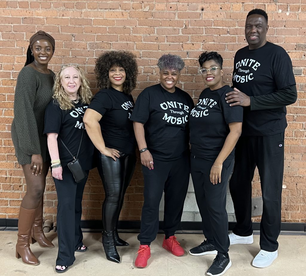 photo of six adults standing in front of a brick wall, smiling and posing for the camera; all but one is wearing a black teeshirt that says 'Unite through Music'