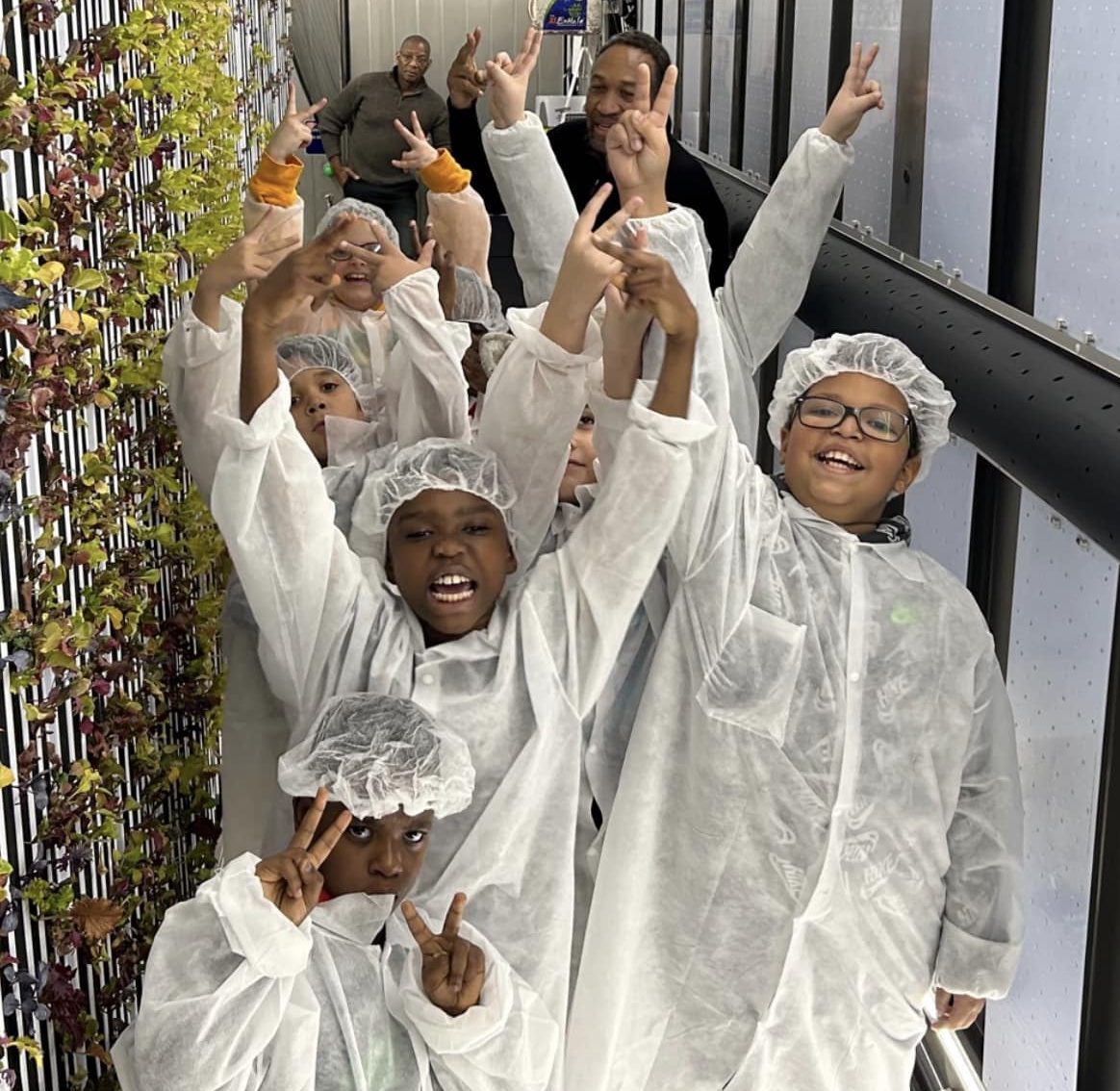 photo of a group of kids all wearing whit clothing covers and hair covers standing in a hallway beside a vertical garden, all looking at the camera, posing and smiling with a couple adults in the background